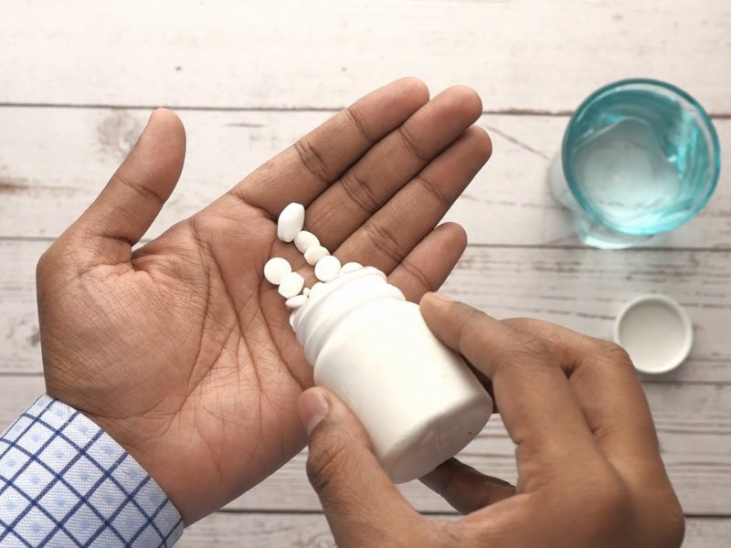 Čovek sipa lekove iz bele tubice na svoj dlan, pored njega je čaša vode i poklopac od tube u kojoj su lekovi a pozadina je neutralna