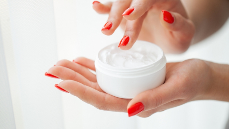 Ruke sa crvenim noktima drže otvorenu teglicu Pantenol kreme spremnu za nanošenje