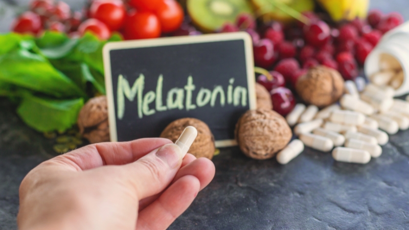 Ruka drži kapsulu melatonina ispred table sa natpisom i hrane poput orašastih plodova i voća
