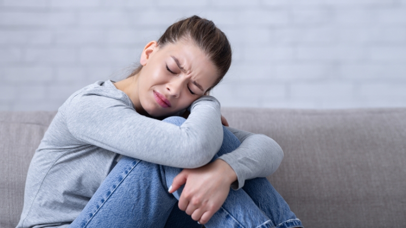 Žena sedi sklupčana na kauču sa vidljivim bolom i iscrpljenošću na licu