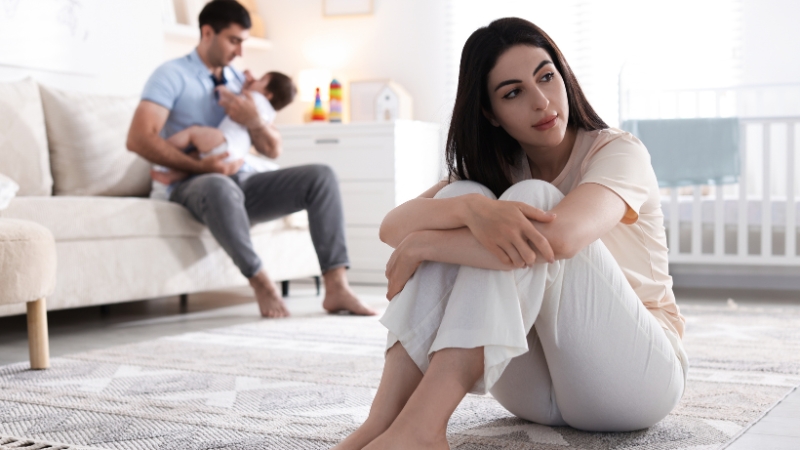 Majka sedi na podu zamišljena i povučena, dok otac u pozadini drži bebu i brine o njoj