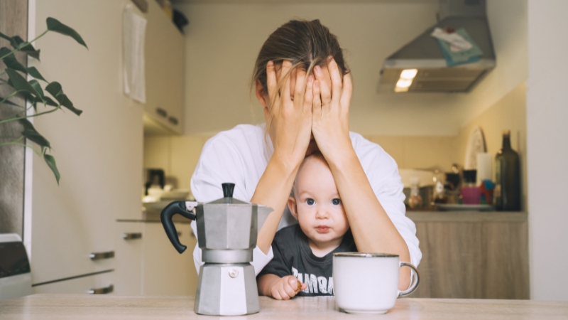 Majka sedi za stolom sa bebom, drži ruke preko lica dok deluje iscrpljeno i preopterećeno