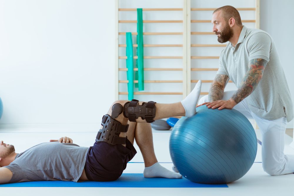 Fizioterapeut pomaže muškarcu sa ortozom na kolenu da izvodi vežbu za jačanje mišića nogu uz pomoć velike plave pilates lopte